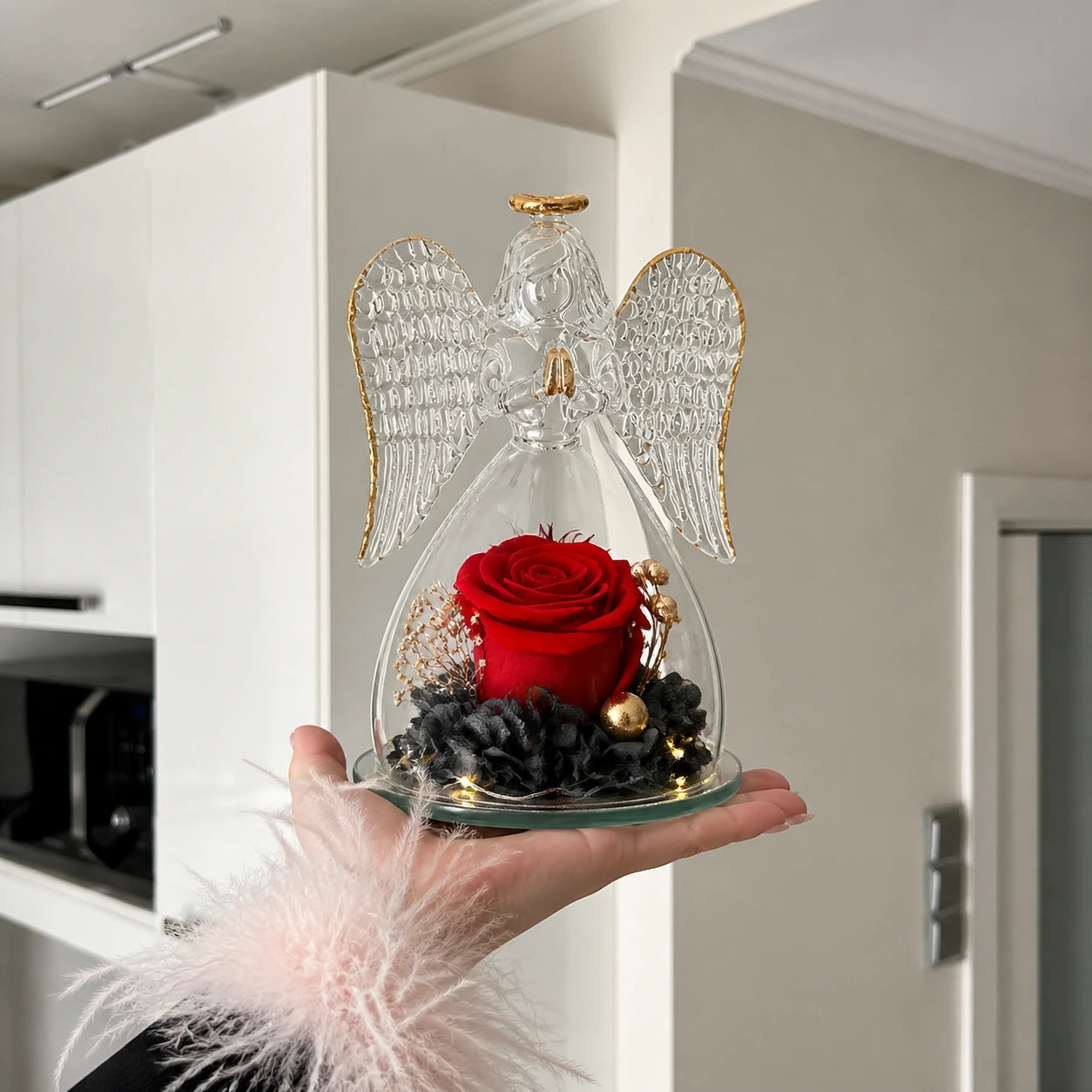Hand hält einen kleinen Glasengel mit einer roten Rose in einer hellen Küche